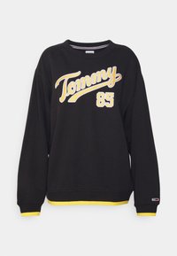 Sweatshirt negro de manga larga, con detalles amarillos en el dobladillo y los puños, que presenta texto bordado en blanco y amarillo "Tommy 85" en el frente.