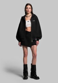 Felpa nera oversize con zip e grandi tasche, abbinata a un top bianco corto e a una gonna nera asimmetrica. Styling con stivali neri fino al ginocchio.