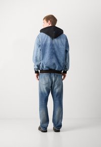 Chaqueta de mezclilla con capucha negra, corte suelto, puños y cintura de canalé oscuros, jeans azul claro con diseño deslavado, aspecto usado, zapatos negros.