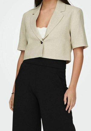 Vrouw die een beige cropped blazer draagt over een wit topje en hoge taille wijde zwarte broek tegen een effen achtergrond.