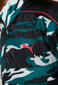 Lätt, svart och teal kamouflage vindjacka med vita accenter och en röd Puma-logotyp; har en halvdragkedja framtill och en slät yta.