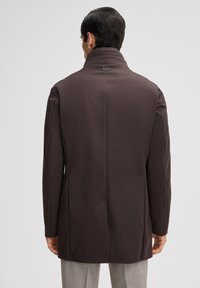 Bruine op maat gemaakte jas met een hoge kraag, gladde textuur, een rugvent en een klein zwart label aan de nek. Draagt over grijze broek.