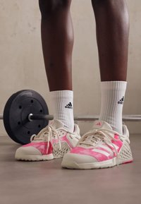 Pantofi sport albi cu accente roz, partea superioară din plasă texturată și talpă din cauciuc gri. Purtați cu șosete albe cu textură, care au branding negru.