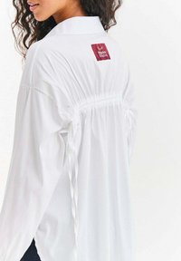 Biała bluzka z długimi rękawami, z marszczonym tyłem ozdobionym wstążkami. Na górze pleców widoczna metka z napisem "Nako Studio".