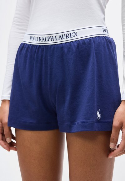 Γυναίκα που φοράει σορτς Ralph Lauren σε ναυτικό μπλε με λευκή ελαστική μέση και κεντημένο λογότυπο στο αριστερό πόδι, συνδυασμένα με λευκό τοπ μακρυμάνικο.