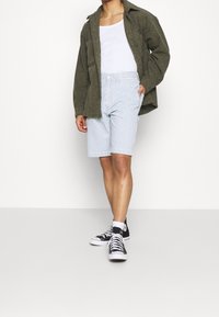 Person som bär en olivgrön jacka, vit linne, ljust blå och vitrandiga knälånga shorts, vita strumpor och svarta högkängor.