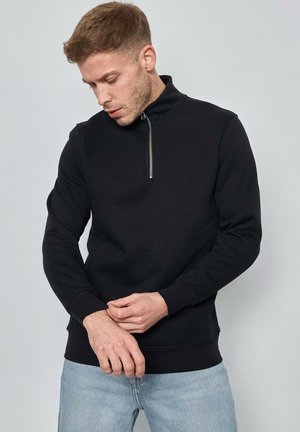 Sweat-shirt zippé noir avec des manches longues et un col haut. Le tissu semble lisse. Assorti à un jean bleu clair. Design minimaliste.