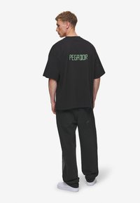 Sort oversized t-shirt med "PEGADOR" i grønne bogstaver på ryggen, sammen med sorte denim jeans og hvide sneakers.