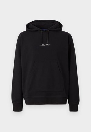 Meesmodell kannab musta A-COLD-WALL kapuutsiga pusa ja lühikesi pükse, hoides suurt halli kotti, seisab ühevärvilise heleda tausta ees.