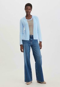 Vaaleansininen neuletrikoo, jossa on pitkät hihat, päällä vaaleanbeige paita, yhdistettynä leveisiin sinisiin farkkuihin ja vaaleanbeigeihin kenkiin.