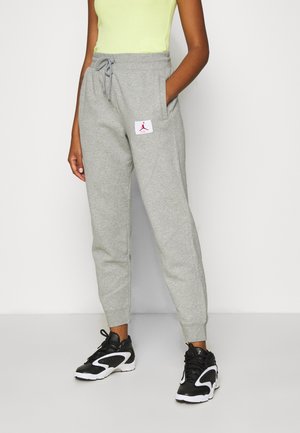 Person iført lysegrå træningsbukser med et hvidt og rødt logopatch og sorte og hvide sports-sneakers på en ensfarvet baggrund.