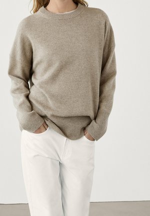 Personne portant un pull en maille beige avec poignets et bas côtelés, légèrement rentré dans un pantalon blanc, les mains dans les poches, sur un fond uni.