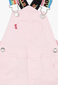 Gli overall in denim rosa presentano un dettaglio di cucitura a forma di cuore, bottoni a pressione argentati e bretelle regolabili a strisce arcobaleno.