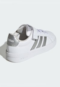 Bílá teniska s hladkým koženým svrškem, stříbrnými doplňky, třemi kovovými pruhy a páskem na suchý zip. Texturovaná podrážka a polstrovaný límec.