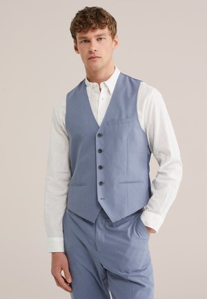 Joven vestido con chaleco azul claro y pantalones a juego sobre una camisa blanca de botones, de pie con una mano en el bolsillo.