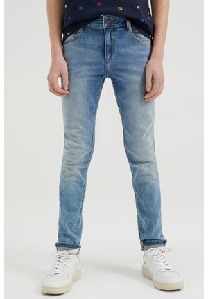 Person trägt hellblaue Slim-Fit-Jeans und weiße Sneaker und steht vor einem einfarbigen hellen Hintergrund.