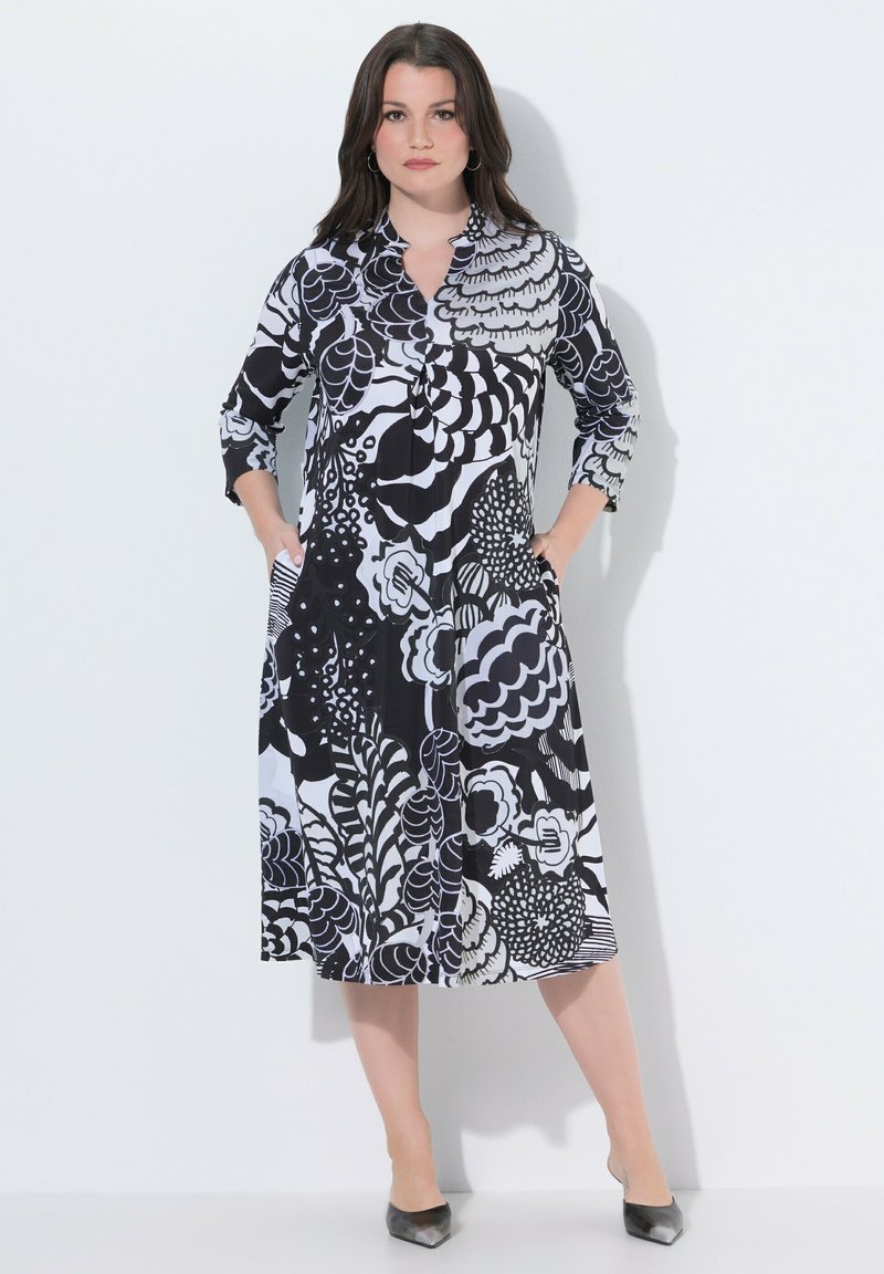 Kvinde iført en sort og hvid mønstret midikjole med trekvartærmer, stående med hænderne i lommerne mod en ensfarvet baggrund.