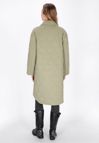 Quilted, lang olivengrøn frakke med krave, som har et diamantmønster og en rund kant. Sammen med sorte knæhøje støvler.