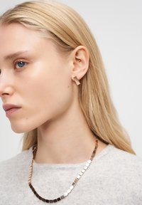 Sidosprofil av en kvinna med blont hår som bär fyrkantiga guldörhängen och ett matchande guldhalsband med fyrkantiga länkar mot en vit bakgrund.