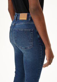 Dunkelblaue Jeans aus Denim mit hohem Bund, versehen mit einem hellbraunen Lederpatch auf der Rückseite und klassischen Gesäßtaschen mit genähten Details.
