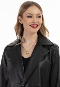 Schwarze Lederjacke mit großem Kragen, kombiniert mit einer goldenen Kette, die einen runden Anhänger mit strukturierten Details trägt.