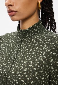 Κοντινό πλάνο ενός ατό�μου που φοράει μια ελαιόλαδη πράσινη φλοράλ μπλούζα με φουσκωτό γιακά, χρυσά σκουλαρίκια κρίκοι, ρινικό και ρινίδιο σκουλαρίκι, και πλεξούδες.