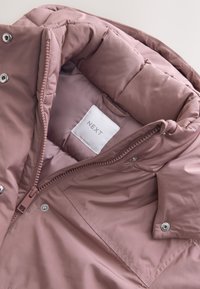 Veste matelassée mauve clair avec un col montant, partiellement zippée, montrant une étiquette de marque blanche "NEXT" à l'intérieur de la zone du cou.