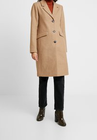 Cappotto in misto lana color cammello con due tasche laterali, design a un petto e bottoni neri, abbinato a jeans neri e stivali con tacco lucidi.