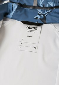 Tessuto bianco con una texture morbida, caratterizzato da un rivestimento a fantasia blu e grigia con motivi di cervi e un'etichetta di cura.