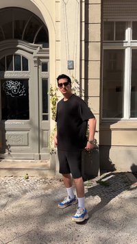 Άνδρας με μαύρο μπλουζάκι, μαύρα σορτς, γυαλιά ηλίου και αθλητικά παπούτσια στέκεται σε λιθόστρωτο δρόμο δίπλα σε μπεζ κτίριο με αψιδωτή πόρτα και παράθυρο.