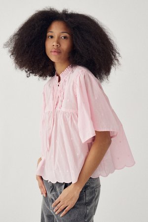 Młoda kobieta o bujnych kręconych włosach, ubrana w luźną, jasnoróżową bluzkę i szare dżinsy, stojąca na tle jednolitego tła.