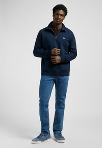 Veste en polaire bleu marine zippée avec un col haut et un logo. Portée avec un jean en denim bleu et des baskets bleu clair. Texture lisse et design décontracté.