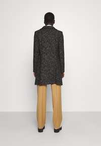 Svart och vit texturerad kappa med en lång silhuett, med en spetsig krage, tillsammans med beige vida byxor och svarta skor.