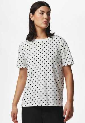 Femme aux cheveux foncés portant une chemise blanche à manches courtes couverte de pois noirs régulièrement espacés, associée à un pantalon noir, légèrement de profil.