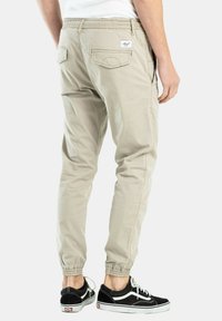 Pantalones jogger beige con cintura elástica, dos bolsillos traseros y puños acanalados, combinados con zapatillas negras. Textura de tela suave.