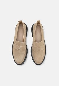Beige zamšas slip-on apavi ar melnām gumijas zolēm, ar joslu pāri pēdas vidusdaļai, kas rotāta ar iepresētu zīmolu un gludu iekšējo oderi.