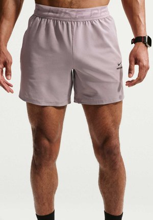 Mand iført lyse lilla Nike Pro-shorts og et sort armbåndsur, står med armene afslappet ned langs siderne mod en ensfarvet baggrund.