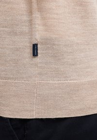 Maglione di maglia beige con una finitura texture, dotato di un'etichetta blu scuro sul lato e orlo a coste, indossato sopra pantaloni scuri.