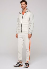 Lys grå polstret jakke med hætte og sort lynlås, parret med hvide joggere med en orange stripe og grå sportsko.