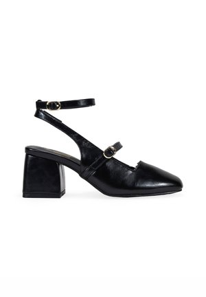 Sandales noires en cuir à talon carré avec un bout pointu, double bride de cheville avec boucles en ton or, et une texture lisse.