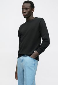 Mladý muž v čiernej košeli s dlhými rukávmi a svetlomodrých džínoch, stojaci s jednou rukou vo vrecku pred čistým bielym pozadím.