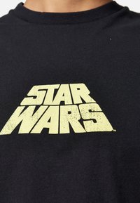 Svart bomullströja med en stor, sliten gul "STAR WARS"-logotyp i mitten. Rund halsringning och korta ärmar.