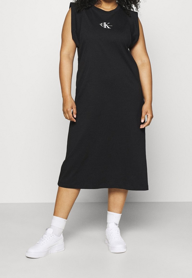 Czarna sukienka bez rękawów wykonana z bawełny, do kolan, z subtelnym logo na piersi. Połączona z białymi trampkami i skarpetkami za kostkę.