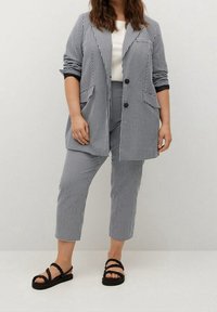 Czarny i biały garnitur w kratkę z dopasowaną marynarką i spodniami do kostek, w połączeniu z czarnymi sandałami na cienkich paskach. Gładka struktura materiału.