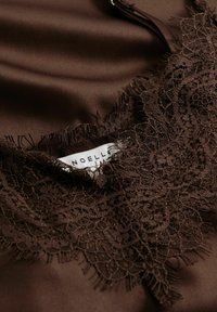 Pruun satiinkangas, millel on pitsist ääris keerukate lillemustrite ja narmastega, ning ka silt nimega "NOELLA".