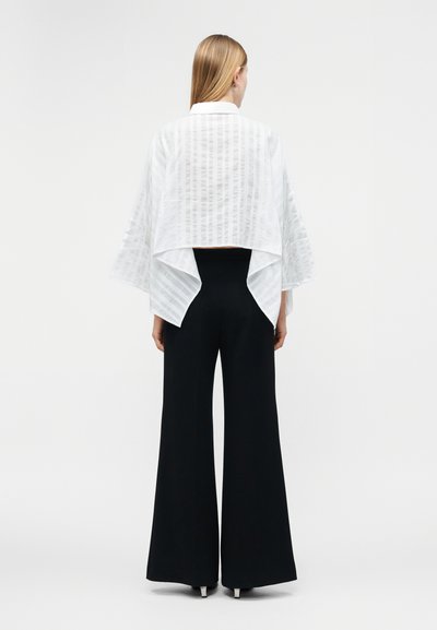 Žena so svetlými rovnými vlasmi, ktorá má na sebe biely voľný top so širokými rukávmi a čierne nohavice s vysokým pásom a širokými nohavicami, obrátená chrbtom.