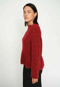 Maglione rosso chunky con un collo alto, maniche lunghe e un dettaglio peplum all'orlo. Realizzato in un tessuto morbido e strutturato.