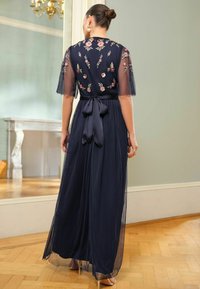 Marineblaues Abendkleid mit floralem Stickereien, durchsichtigen Ärmeln und gebundenem Taillenbereich. Langer, fließender Rock und weiche Textur. Klarer Rückansicht.