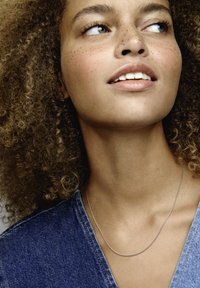 Collar de cadena de plata llevado sobre una camiseta de mezclilla. El collar tiene una textura suave y se adapta de manera plana sobre la piel. La modelo tiene el cabello rizado.