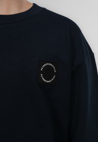 Navyblauwe sweatshirt met een ronde halslijn, oversized pasvorm en een zwart vierkant patch met witte tekst en logo op de linkerkant van de borst.